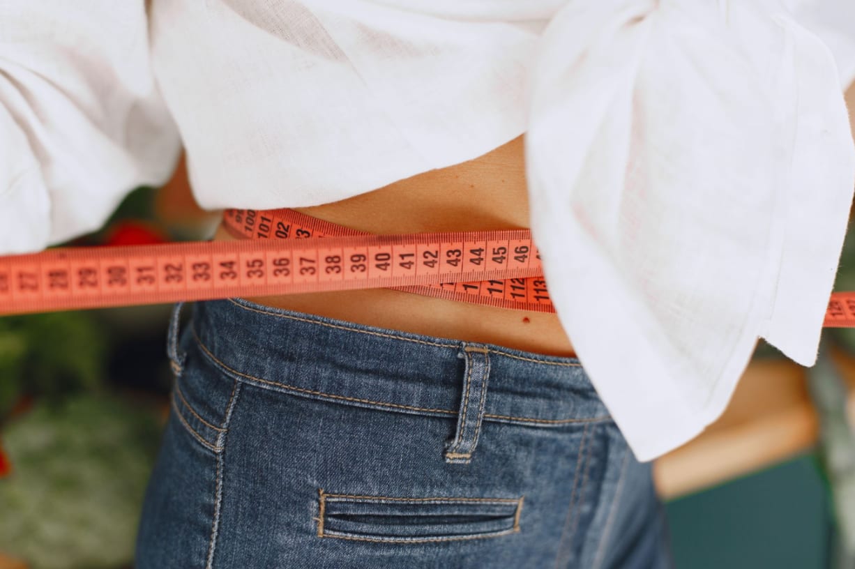 A tape measure around a woman's waist over loose jeans, illustrating how significant or rapid weight loss reduces facial fat and can make the eyes look more hollow